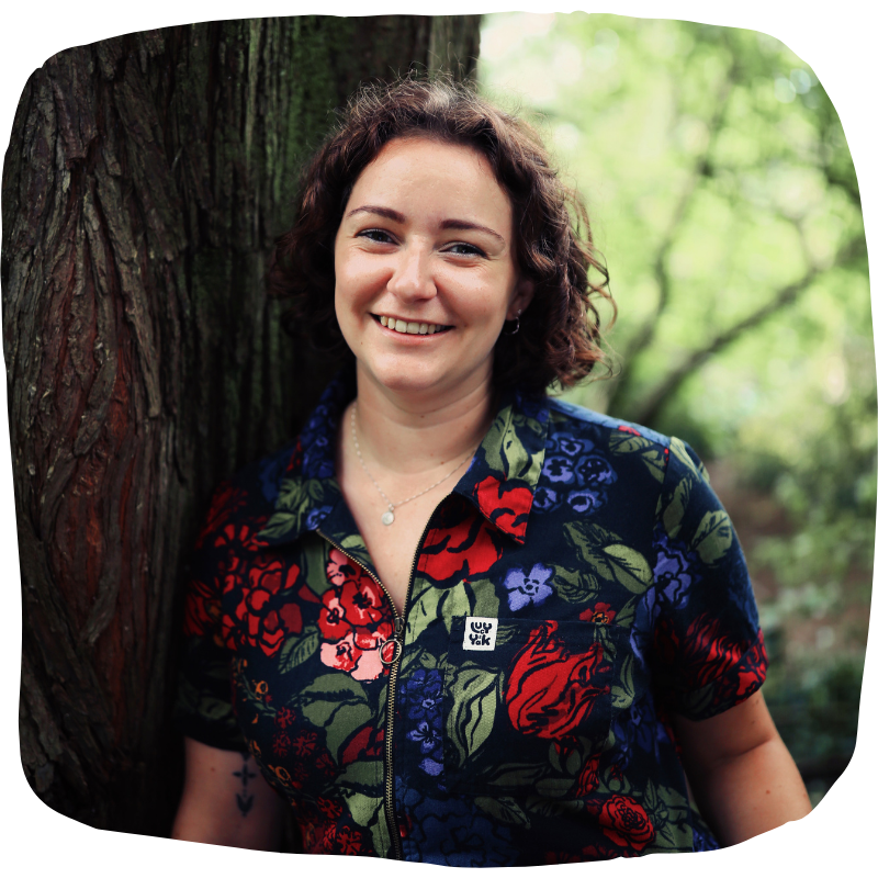 Close-up photo of Laura leaning against a tree. She is wearing a floral patterned jumpsuit with a collar and a zip. She is looking straight at the camera with a big smile.