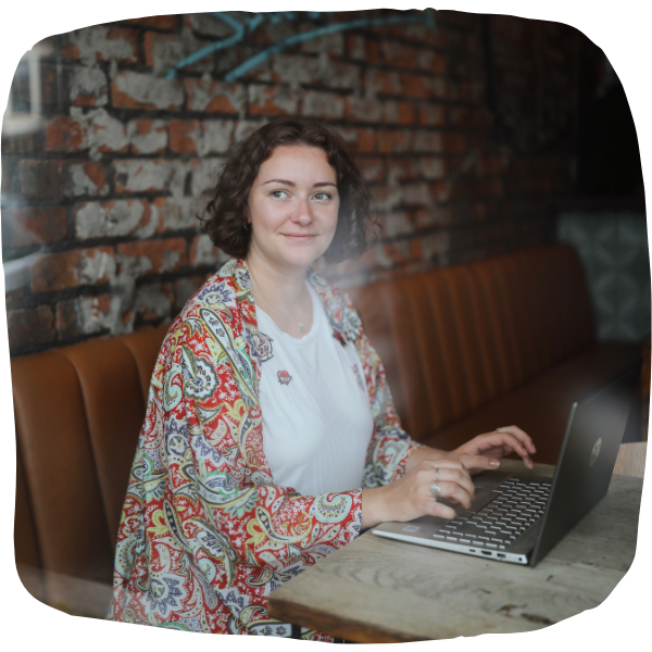 Photo of Laura sitting on a leather bench in front of a wooden table, typing on a laptop. She is wearing a white t-shirt with a red, paisley kimono. She is looking out of the window, away from the camera and has a closed smile on her face.