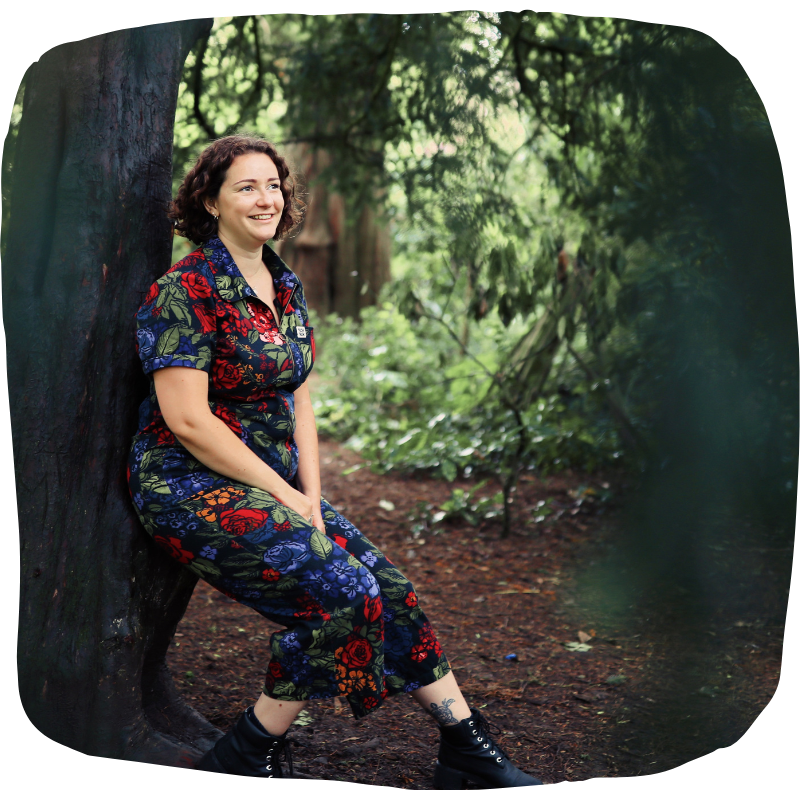 Full-length photo of Laura sitting with her back against a tree. She is wearing a floral patterned jumpsuit with a collar and a zip and black lace-up boots. She is looking up and to the right of the camera with a big smile on her face.