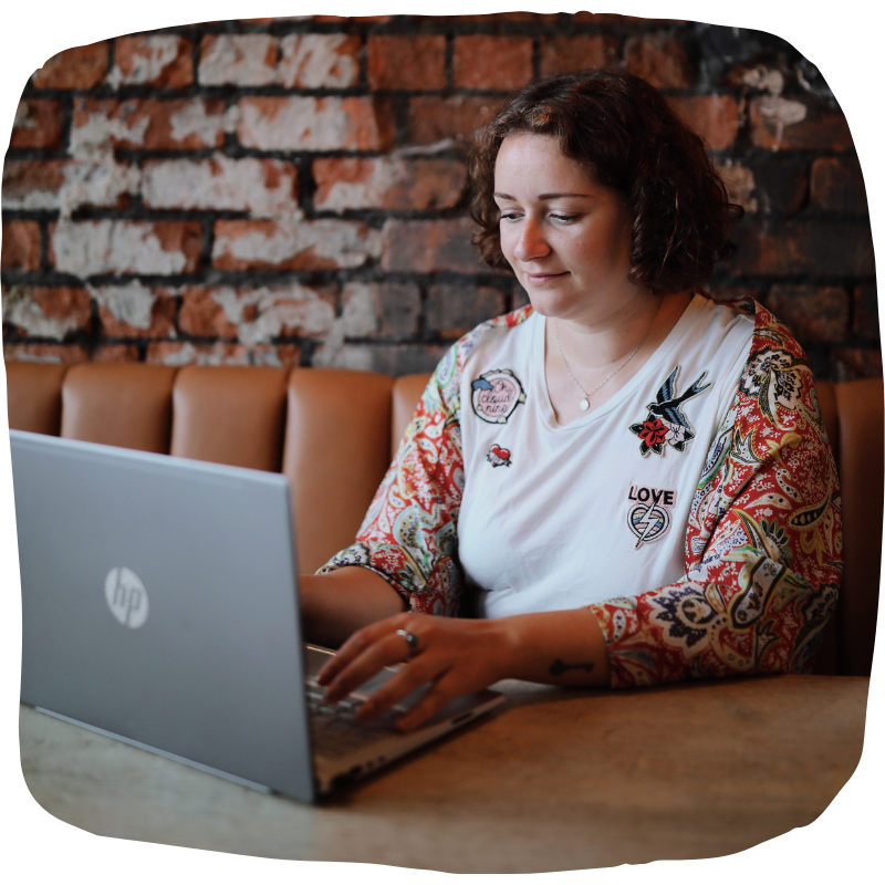 Photo of Laura sitting on a leather bench in front of a red brick wall, typing on a laptop. She is wearing a white t-shirt with a red, paisley kimono. She is looking at her laptop screen.